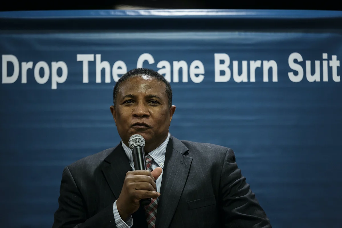 A man dressed in a suit stands in front of a blue banner reading, "Drop The Cane Burn Suit."