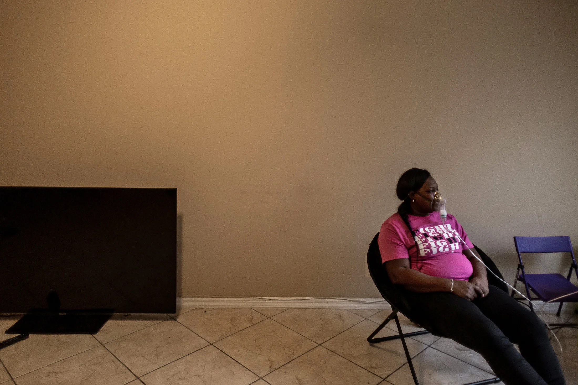 A woman sits on the right side of the photo, wearing the same nebulizer mask from the previous image. She's in front of a blank wall, and a television sits on the floor on the left side of the photo. The woman is wearing a pink T-shirt reading, "FIGHT FIGHT FIGHT FIGHT FIGHT."