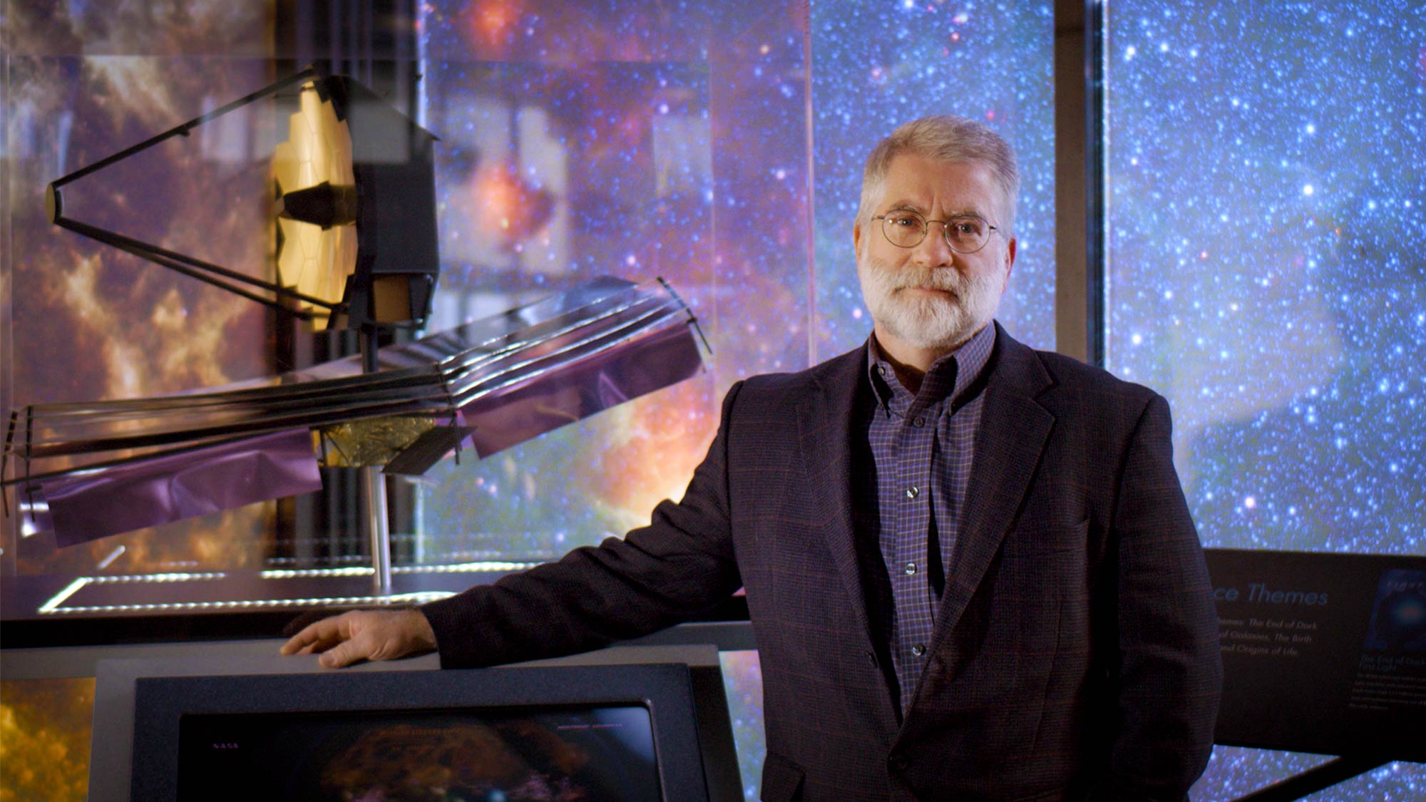 This image shows Michael Menzel standing in front of a starry backdrop and with a model of the JWST.